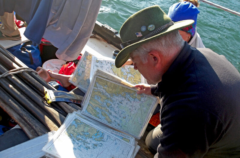 In the image, a man wearing a green hat is looking at maps on a boat. He appears to be studying them intently. Other people are visible in the background, and the presence of water suggests they are at sea. The scene conveys a sense of navigation or exploration.